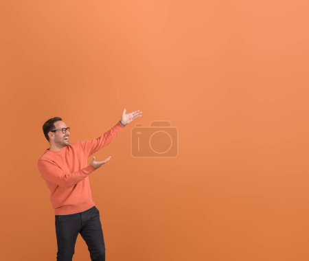 Ekstatischer Jungunternehmer lacht und präsentiert neues Produkt auf leerem Raum vor orangefarbenem Hintergrund