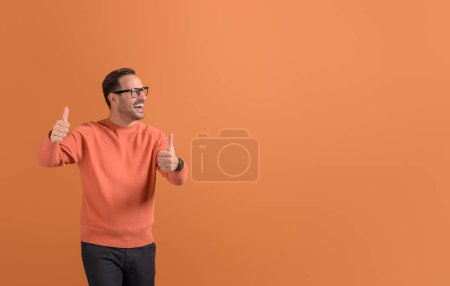 Succès jeune homme d'affaires montrant pouce vers le haut signe et regardant loin joyeusement sur fond orange