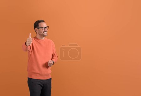 Porträt eines glücklichen männlichen Unternehmers, der auf orangefarbenem Hintergrund ein Zeichen mit hochgereckten Daumen zeigt und wegschaut