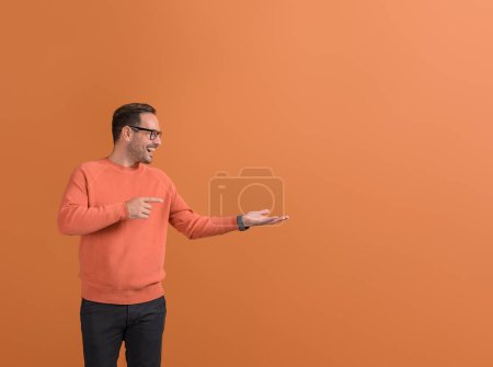 Ekstatischer Verkäufer zeigt auf leere Handfläche und demonstriert neues Produkt auf orangefarbenem Hintergrund
