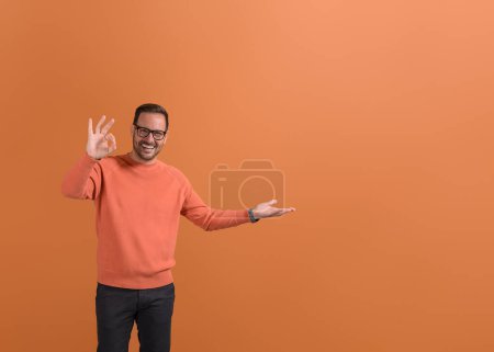Portrait of smiling young businessman showing empty palm and OK sign on isolated orange background