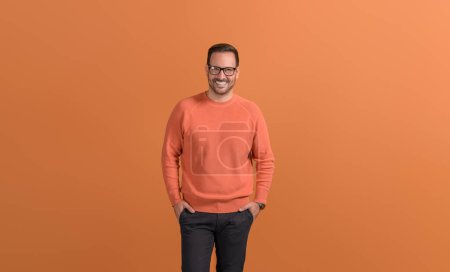 Portrait of smiling male freelancer with hands in pockets standing confidently on orange background