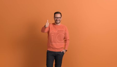 Junger erfolgreicher Geschäftsmann mit Brille lächelt und zeigt Daumenhoch-Zeichen auf orangefarbenem Hintergrund