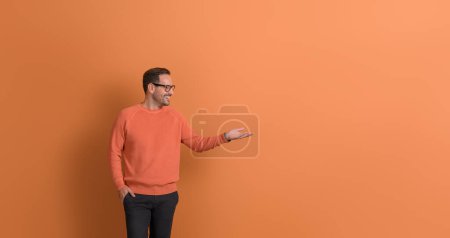 Photo for Happy male promoter with hand in pocket looking at empty palm while advertising on orange background - Royalty Free Image