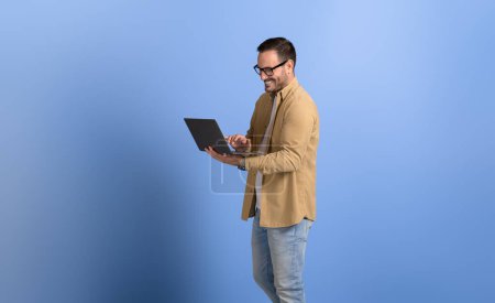 Homme gestionnaire de lunettes souriant et travaillant en ligne sur un ordinateur sans fil sur fond bleu