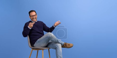 Young businessman laughing and talking on speakerphone while sitting on chair over blue background