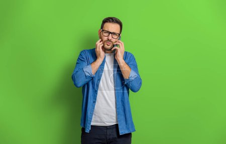 Porträt eines jungen Mannes mit verwirrtem Gesichtsausdruck, der vor grünem Hintergrund mit dem Handy spricht