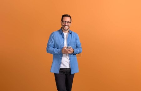 Porträt eines jungen, gut aussehenden Geschäftsmannes, der über sein Handy auf orangefarbenem Hintergrund lächelt und Nachrichten verschickt
