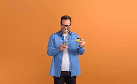 Smiling young man buying online over smart phone and paying through credit card on orange background