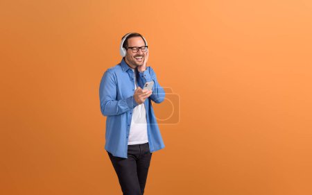 Glücklicher junger Mann mit geschlossenen Augen, der Musik über Kopfhörer und Telefon auf orangefarbenem Hintergrund genießt