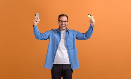 Foto de Feliz cliente masculino con tarjeta de crédito y teléfono celular levantando brazos y gritando sobre fondo naranja - Imagen libre de derechos
