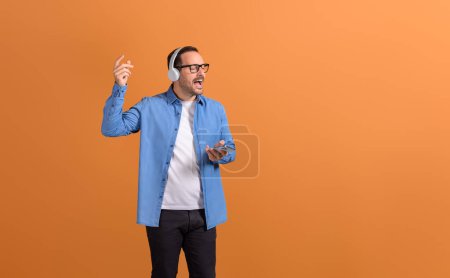Young man with eyes closed singing song and listening music over headphones on orange background