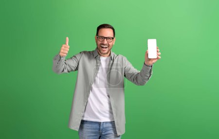 Retrato de un joven hombre de negocios mostrando el signo de pulgares hacia arriba y la pantalla del teléfono móvil sobre fondo verde