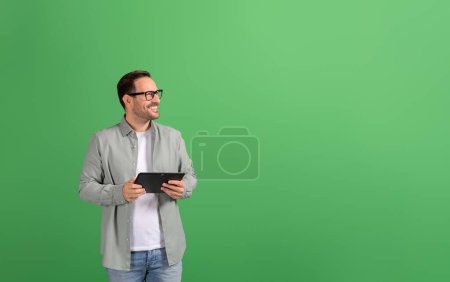 Erfolgreicher Geschäftsmann mit digitalem Tablet lächelt und schaut nachdenklich auf grünem Hintergrund weg