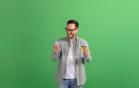Happy young businessman using credit card to make online payment on smartphone over green background