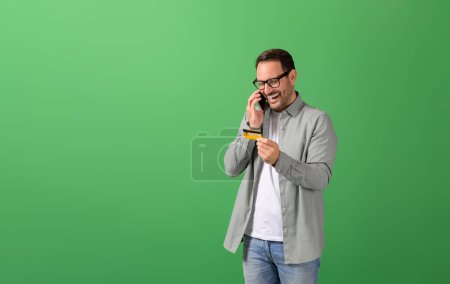 Fröhlicher junger Mann teilt Kreditkartendaten mit, während er auf dem Smartphone vor grünem Hintergrund spricht
