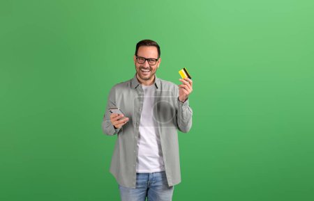 Portrait of satisfied male customer suggesting mobile payment with credit card on green background