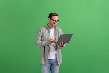 Porträt eines glücklichen, gutaussehenden Unternehmers, der vor grünem Hintergrund E-Mails online über Laptop checkt