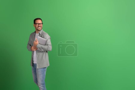 Positive businessman holding laptop and looking away thoughtfully while standing on green background