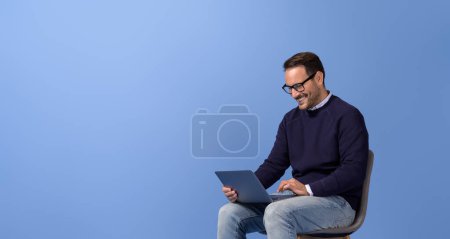 Homme d'affaires souriant faisant des recherches en ligne sur ordinateur sans fil sur chaise sur fond bleu