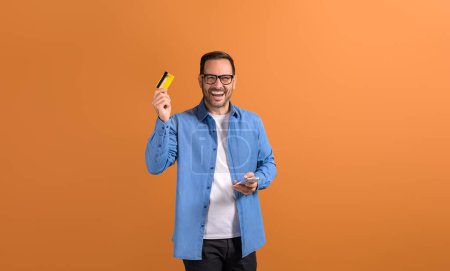 Fröhlicher junger Verkäufer lacht und zeigt Kreditkarte, während er Smartphone auf orangefarbenem Hintergrund benutzt