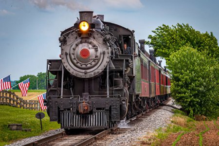 Photo for View of a Restored Steam Train Approaching Head-On With American Flags Waving on a Fence on a Spring Day - Royalty Free Image