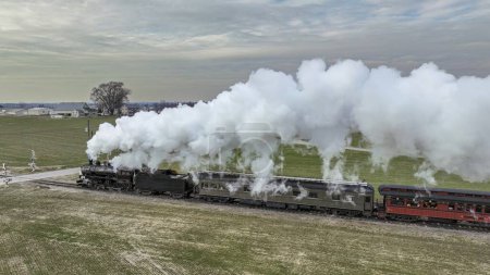 Foto de Una vista aérea lateral de un tren de pasajeros de vapor acercándose, viajando a través de tierras agrícolas abiertas, soplando mucho humo blanco, en un día de invierno - Imagen libre de derechos