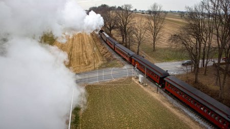 Photo for A Behind Aerial View of a Steam Passenger Train Approaching, Traveling Thru Open Farmlands, Blowing Lots of White Smoke, on a Winter Day - Royalty Free Image