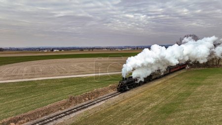Photo for An Aerial View of a Steam Passenger Train Approaching, Traveling Thru Open Farmlands, Blowing Lots of White Smoke, on a Winter Day - Royalty Free Image