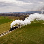 An Aerial View of a Steam Passenger Train Approaching, Traveling Thru Open Farmlands, Blowing Lots of White Smoke, on a Winter Day