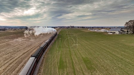 Photo for A Behind Aerial View of a Steam Passenger Train Approaching, Traveling Thru Open Farmlands, Blowing Lots of White Smoke, on a Winter Day - Royalty Free Image