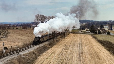 Photo for A Drone View of a Steam Locomotive Approaching Traveling Thru Fields and Meadows, Blowing White Smoke on a Winter Day - Royalty Free Image