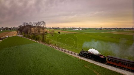 Foto de Un poco detrás de la vista aérea de un antiguo tren de vapor de pasajeros que viaja en una sola vía a través de la campiña y las tierras de cultivo, en un día de primavera - Imagen libre de derechos