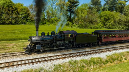 Photo for An Aerial View from the Side of An Antique Steam Locomotive and Passenger Coach Stopped and Blowing Smoke and Steam, While Waiting for Passengers to Board on a Sunny Spring Day - Royalty Free Image