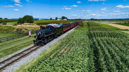 Photo for A Drone Front View of a Steam Passenger Train Approaching, Traveling Thru Farmlands and Passing a Fence With An America Flag on ti, on a Sunny Summer Day - Royalty Free Image