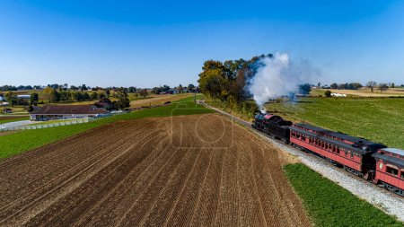 Téléchargez les photos : Une vue de drone de derrière et au-dessus d'un train de voyageurs à vapeur voyageant à travers les terres agricoles et soufflant de la fumée un jour d'automne ensoleillé - en image libre de droit