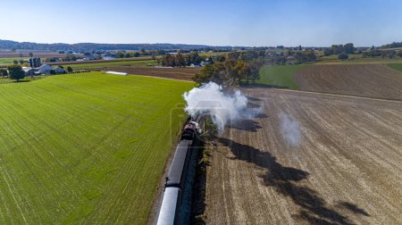 Photo for A Drone View From Behind and Above of a Steam Passenger Train Traveling Thru Farmlands and Blowing Smoke on a Sunny Fall Day - Royalty Free Image