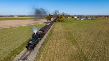 Foto de Una vista del dron desde arriba y delante de un tren de pasajeros de vapor que viaja a través de tierras de cultivo y que sopla humo en un día soleado de otoño - Imagen libre de derechos