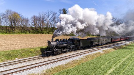 Téléchargez les photos : Un train de voyageurs à vapeur antique voyageant à travers les terres agricoles, avec des cultures plantées, soufflant de la fumée un jour de printemps ensoleillé - en image libre de droit