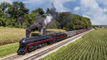 Foto de Una vista frontal aérea de un tren de pasajeros de vapor antiguo restaurado, viajando a través del campo, pasando campos de maíz y soplando humo en un soleado día de verano - Imagen libre de derechos