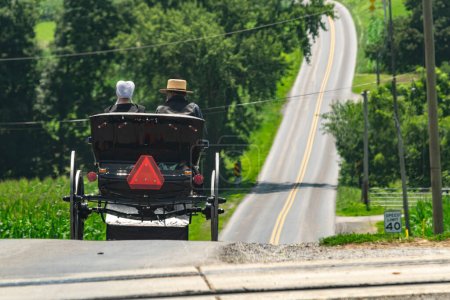 Die Rückansicht eines Amish-Paares in offenem Pferd und Buggy, das an einem sonnigen Sommertag eine Landstraße hinunterfährt