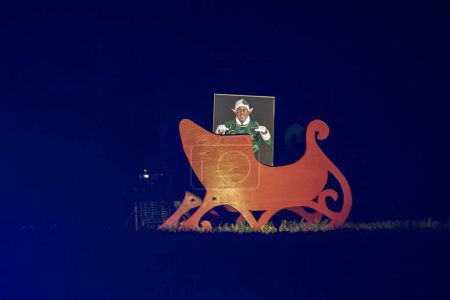 Nighttime View Of A Large Wooden Cut-Out Of Santas Sleigh With A Person Posing As An Elf Through A Face-In-Hole Board, Creating A Festive Photo Opportunity.