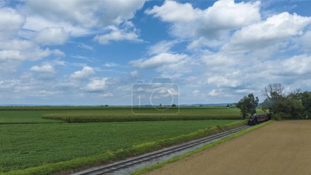 Railroad Tracks Curve Gently Through A Lush Agricultural Landscape With Fields Of Various Shades Of Green Under A Wide Sky With Broken Clouds.