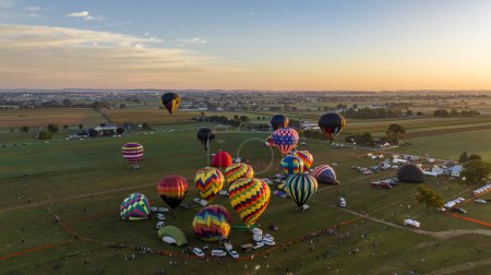 Foto de Una vista aérea del cúmulo de globos de aire caliente coloridos que se preparan para el vuelo al amanecer con espectadores y vehículos reunidos en un campo rural. - Imagen libre de derechos