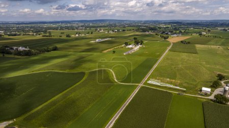 Photo for Expansive Aerial View Of Rolling Green Agricultural Fields, Farm Buildings, And A Long Country Road Under A Cloudy Sky. - Royalty Free Image