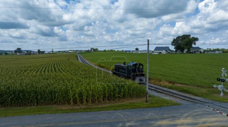 Dark Green Locomotive Moving On Tracks Beside A Cornfield With A Crossing Signal In The Foreground And Farm Buildings In The Distance Under A Dramatic Cloudy Sky.