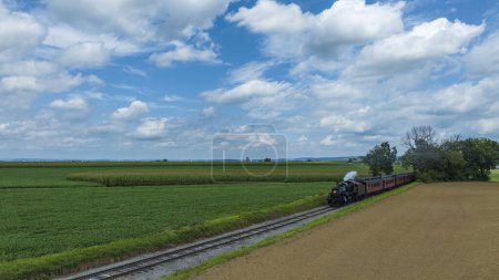 Oldtimer-Dampflokomotive zieht rote Personenwagen neben saftigen Feldern und einem Feldweg unter freiem Himmel mit flauschigen Wolken.