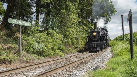 Photo for Black Steam Locomotive Number 89 Blowing Steam On Rural Tracks Next To Carpenters Sign Surrounded By Verdant Foliage. - Royalty Free Image