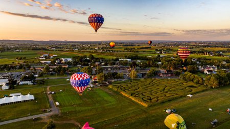 Foto de Una vista aérea de múltiples globos de aire caliente se elevan sobre un paisaje pintoresco con un laberinto de maíz, ciudad y campos durante una puesta de sol de hora dorada. - Imagen libre de derechos