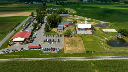 Overhead View Of A Bustling Country Market With A Red Roof, Adjacent To A Farm, Parking Lot Full Of Cars, Railroad Tracks, And Surrounding Green Fields.